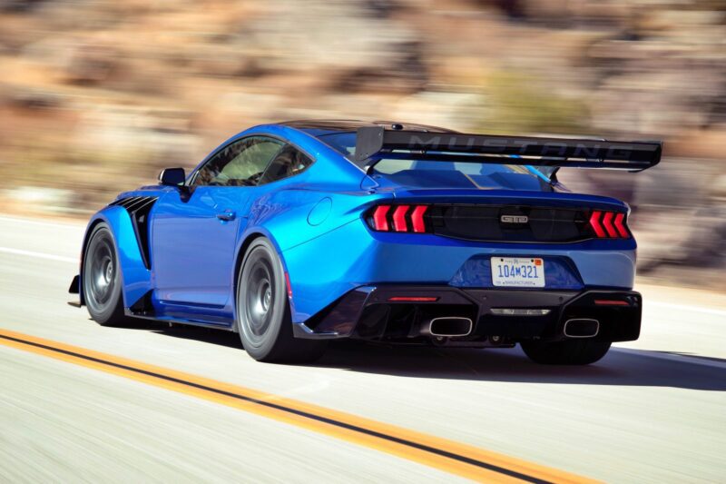 A blue Ford Mustang GTD with a large rear wing drives on a road, blurred rocky scenery behind—capturing the thrill of the First Drive: Six-figure Ford Mustang GTD Boasts Peak Pony Muscle.