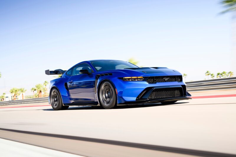 Un auto deportivo azul con un alerón trasero circula por una pista de carreras, con un desenfoque de movimiento en el fondo y palmeras a los costados, capturando la emoción de una primera conducción: un Ford Mustang GTD de seis cifras presume de un músculo de poni de gran tamaño.