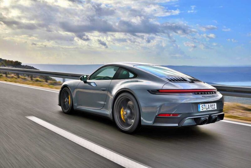 Review: 2025 Porsche 911 Carrera GTS T-Hybrid Electrifies Potent Performance as a silver Porsche 911 sports car cruises along a coastal highway under a partly cloudy sky.
