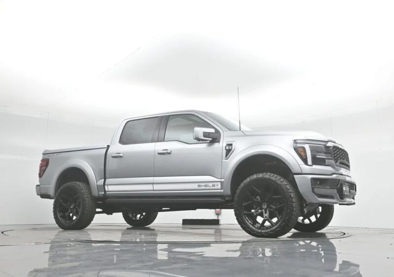 A silver Ford F-150 pickup truck with large black wheels and bold "SHELBY" branding is parked in a white studio setting, showcasing the premium style of Shelby trucks.