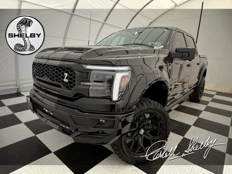A black Shelby F-150 truck is parked indoors on a checkered floor, showcasing the Shelby logo and Carroll Shelby’s signature—a stunning example of Shelby Trucks for sale.
