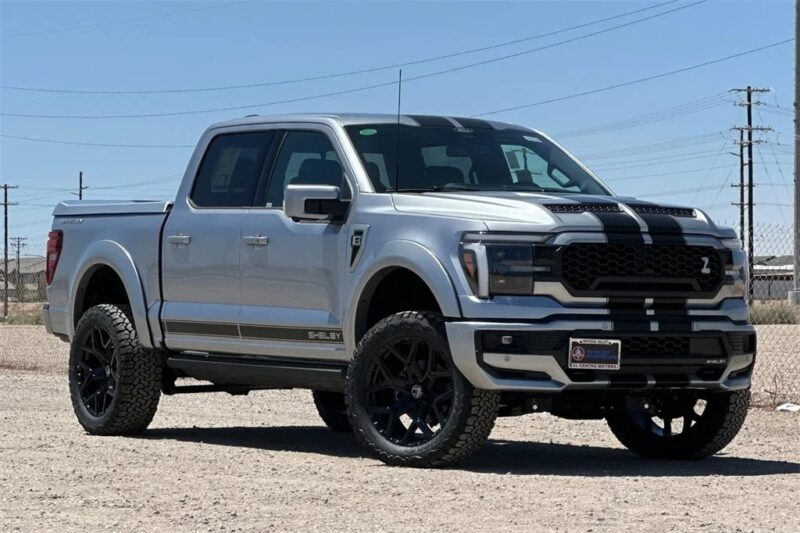 A silver Shelby Ford F-150 pickup truck with black racing stripes is parked on a dirt lot under a clear sky, showcasing the bold styling Shelby Trucks are known for.