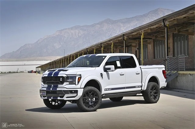 A white Ford F-150 pickup with blue racing stripes, inspired by Shelby Trucks, is parked in front of an industrial building with mountains in the background.