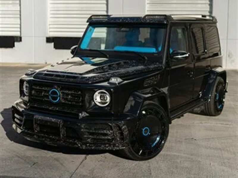 A customized black Mercedes-Benz G-Class luxury car with blue accents is parked on a concrete surface in front of a building with white garage doors.