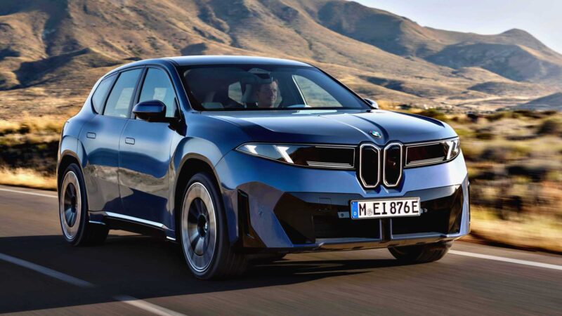 A blue BMW SUV, inspired by the BMW iX3 launching the Neue Klasse era with a 400-mile range and 463 HP, drives on a paved road through a mountainous desert landscape under a clear sky.
