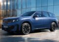 A blue BMW SUV, symbolizing the BMW iX3 launches the Neue Klasse era with 400-mile range and 463 HP, is parked in front of a modern glass building, reflecting sunlight.