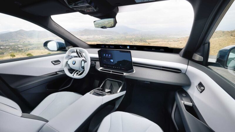 Interior of a modern BMW car featuring a white steering wheel, digital dashboard, central touchscreen, and a panoramic windshield with countryside views—showcasing the innovation behind the BMW iX3 launching the Neue Klasse era with 400-mile range and 463 HP.