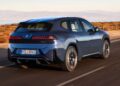 A blue BMW iX electric SUV cruises along a coastal road, mountains and a wooden fence in the background—hinting at the innovation seen as BMW iX3 launches the Neue Klasse era with a 400-mile range and 463 HP.