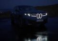A dark SUV with illuminated front grille and headlights is parked on a wet road at night, hinting at the power and innovation of the BMW iX3, which launches the Neue Klasse era with a 400-mile range and 463 HP. Faint city lights glow in the background.