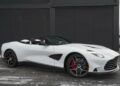 A white Aston Martin convertible performance machine with the top down is parked on a paved surface in front of a matte black wall.