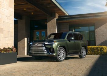 A green Lexus SUV is parked on a stone driveway in front of a modern house with large windows and stone walls.