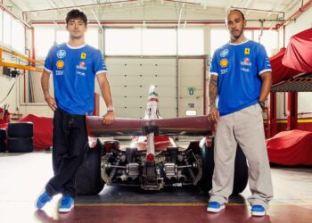 Two men in blue PUMA x Ferrari shirts stand on either side of a Formula 1 car in a garage, with covered vehicles and tires visible in the background.