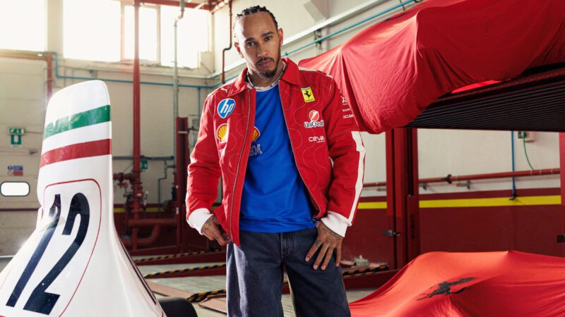 A person wearing a limited-edition PUMA x Ferrari red jacket and blue shirt stands in a garage next to a covered car and a racing kart marked with the number 12.