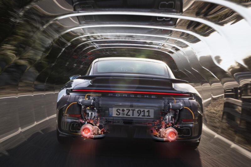 Rear view of the Most Powerful Production 911: 2026 Porsche 911 Turbo S T-Hybrid packs 701HP, showcasing glowing red components and exposed engine parts as it speeds through a tunnel-like blur.