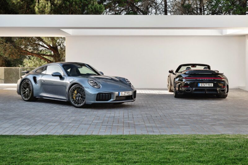 Two silver Porsche 911 Turbo S sports cars, including the most powerful production 911—the 2026 Porsche 911 Turbo S T-Hybrid packing 701HP—are parked on a paved driveway under a modern white canopy, with greenery and trees in the background.