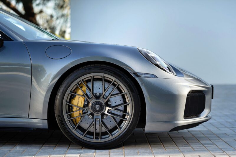 Close-up of a silver sports car’s front left wheel with a large black alloy rim, yellow brake caliper, and drilled brake disc—showcasing the Most Powerful Production 911: the 2026 Porsche 911 Turbo S T-Hybrid packs 701HP.
