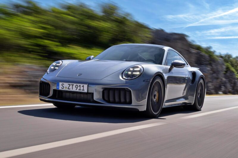 A silver Porsche 911 Turbo S T-Hybrid, the most powerful production 911 with 701HP, speeds down a paved road as blurred greenery and rocks in the background highlight its impressive motion.