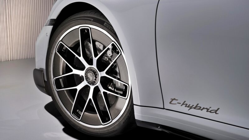 Close-up of a car’s front wheel and fender, featuring a black and silver alloy rim and "t-hybrid" badge on the white body panel—highlighting the Most Powerful Production 911: 2026 Porsche 911 Turbo S T-Hybrid Packs 701HP.