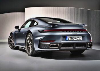A grey Porsche 911 Turbo S, the Most Powerful Production 911: 2026 Porsche 911 Turbo S T-Hybrid Packs 701HP, is shown from the rear at an indoor display, highlighting its taillights, spoiler, rear bumper, and quad exhaust pipes.
