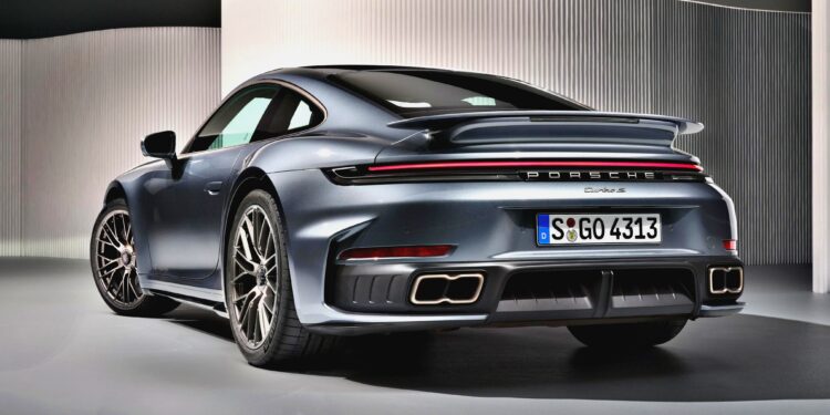A grey Porsche 911 Turbo S, the Most Powerful Production 911: 2026 Porsche 911 Turbo S T-Hybrid Packs 701HP, is shown from the rear at an indoor display, highlighting its taillights, spoiler, rear bumper, and quad exhaust pipes.