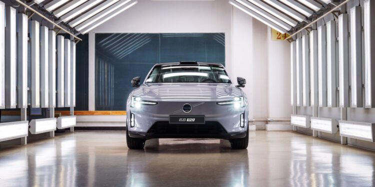 A silver Volvo EX30 electric car is parked indoors under bright overhead lights, facing forward in a modern, industrial setting.