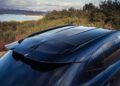 A close-up view of the rear roof and spoiler of a dark-colored 2026 Aston Martin DBX S First Drive: The Best Super SUV Gets Even Better, parked outdoors with trees, hills, and a lake in the background.