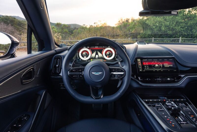 View of a modern luxury car interior from the driver's seat, featuring the 2026 Aston Martin DBX S First Drive: The Best Super SUV Gets Even Better with its digital dashboard, branded steering wheel, and advanced touchscreen infotainment system.