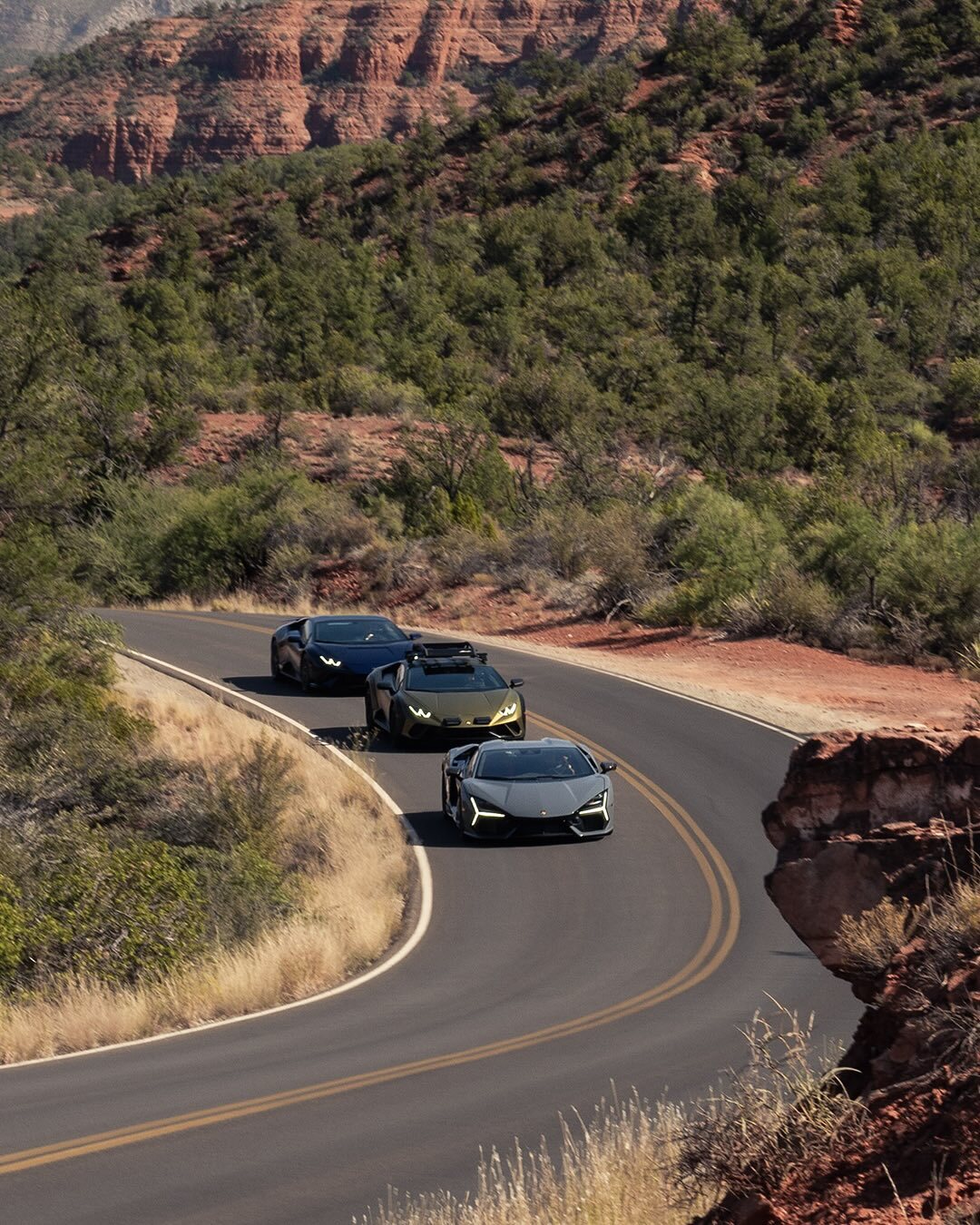 Three sports cars drive along a winding road through a scenic desert landscape near Palm Springs, with red rock formations and greenery—an exhilarating SHEdrives experience.
