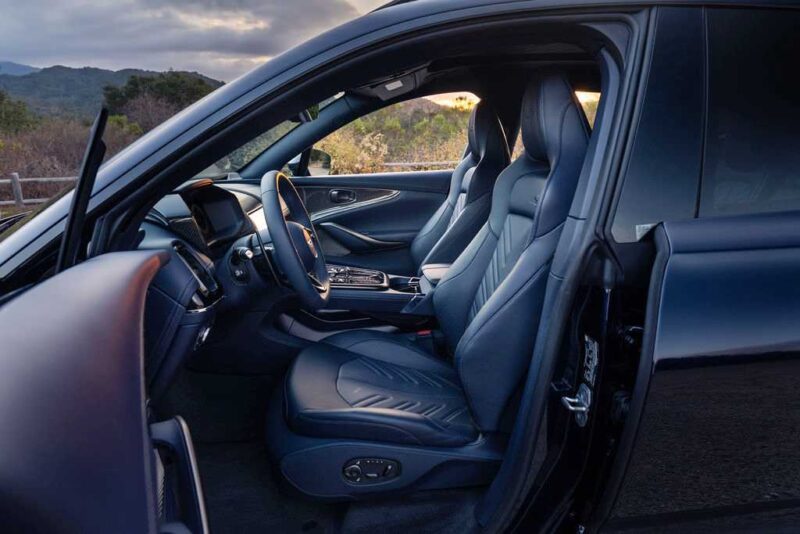 The image shows the interior of a modern car with black leather seats and an open driver-side door, parked outdoors near mountains—evoking the luxury of the 2026 Aston Martin DBX S First Drive: The Best Super SUV Gets Even Better.