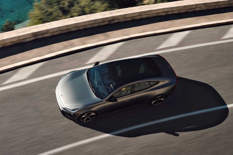 A silver Polestar 5 Debuts at IAA 2025 as Brand’s First True Flagship EV GT sedan drives along a curved road bordered by a stone wall, with greenery visible in the background.