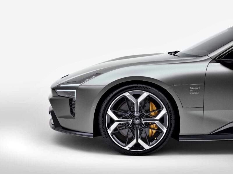 A close-up view of the front left side of a matte gray Polestar 5, the brand’s first true flagship EV GT debuting at IAA 2025, highlights the wheel, brake caliper, and headlight against a white background.
