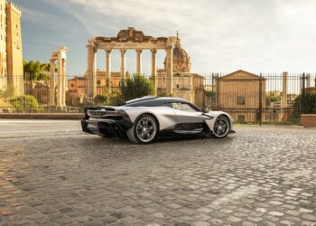 A silver sports car is parked on a cobblestone street in front of ancient Roman ruins under a partly cloudy sky.