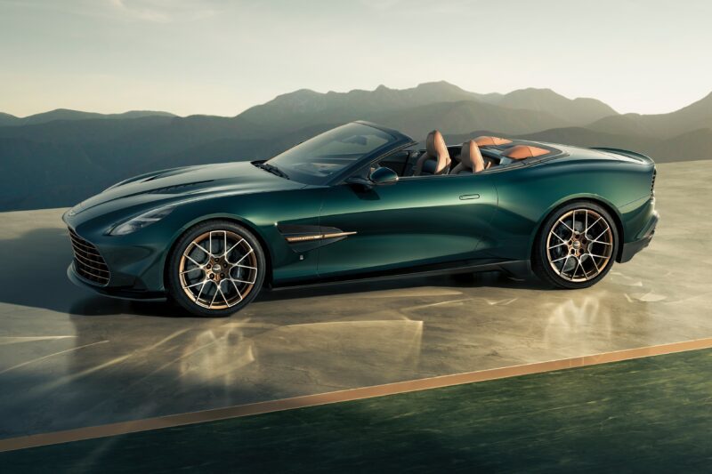 A green convertible sports car, part of the Q by Aston Martin Marks 60 Years of Volante With Ultra-Exclusive Anniversary Editions, is parked on a smooth surface with mountains in the background under clear skies.