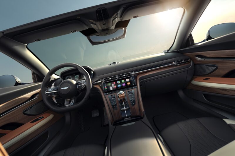 Interior view of a luxury convertible car with wood trim and touchscreen display, celebrating Q by Aston Martin Marks 60 Years of Volante With Ultra-Exclusive Anniversary Editions, featuring a clear sky through the open roof.