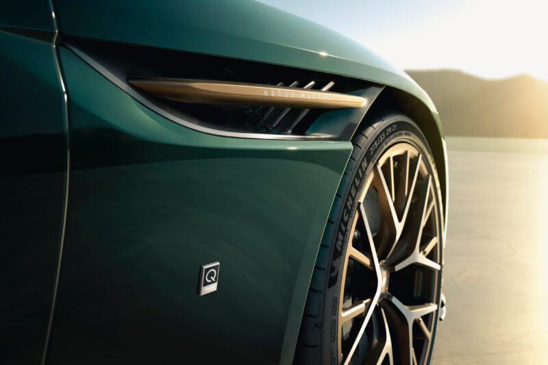 Close-up of a green Aston Martin car’s front fender, wheel, and side vent with sunlight in the background, celebrating Q by Aston Martin Marks 60 Years of Volante With Ultra-Exclusive Anniversary Editions.