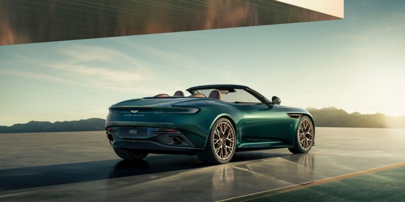 A green Aston Martin convertible sports car, part of the ultra-exclusive Q by Aston Martin Marks 60 Years of Volante Anniversary Editions, is parked on a modern concrete surface with mountains visible in the background under a clear sky.
