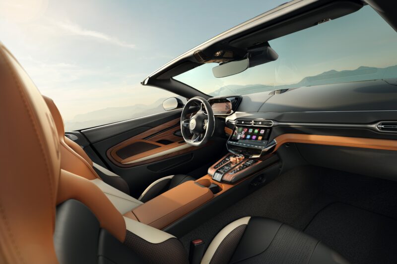 Interior view of a modern convertible car with tan and black leather seats, digital dashboard, and infotainment system—an homage to Q by Aston Martin Marks 60 Years of Volante With Ultra-Exclusive Anniversary Editions—parked top down, overlooking a scenic landscape.