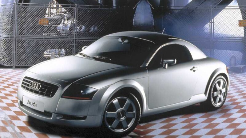 A silver Audi TT coupe is parked indoors on a checkered floor, set against industrial equipment and metal fencing—the perfect backdrop as Audi looks toward its next flagship EV and the anticipated Audi TT successor.