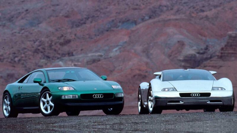 Two Audi concept cars, including the TT Successor and a Flagship EV, stand side by side on a barren landscape with reddish hills in the background.