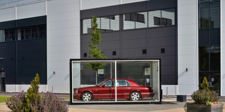 A red Bentley is displayed inside a transparent glass case outside a modern Tech Center, symbolizing the brand's move toward electrification against the building's gray and white exterior walls.