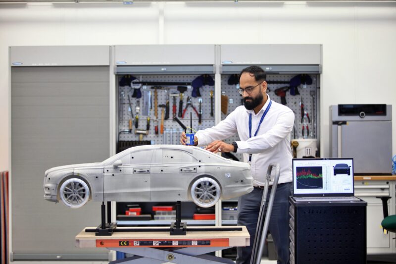 In a Tech Center lab, a man works on a Bentley car model placed on a stand, with tools in the background and a laptop displaying an electrification graph on a nearby table.
