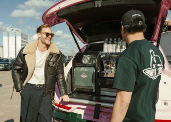 Two men stand by an open Bentley Bentayga trunk, transformed into a stylish Bentayga Coffee Bar with cups and equipment visible, on a sunny day in an urban parking lot.