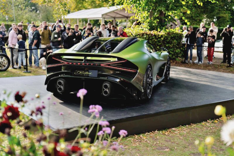 A matte green 1-of-1 Bugatti Brouillard makes its European debut at Wheels Mariënwaerdt, displayed on a raised platform outdoors amid a crowd of onlookers and lush greenery.