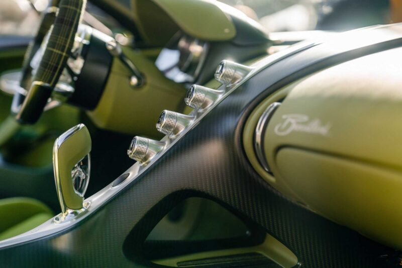 Close-up of a car's interior featuring chrome control knobs, a gear selector, and dashboard with "Bertone" branding—all in green and carbon fiber finish—in the unique 1-of-1 Bugatti Brouillard making its European debut at Wheels Mariënwaerdt.