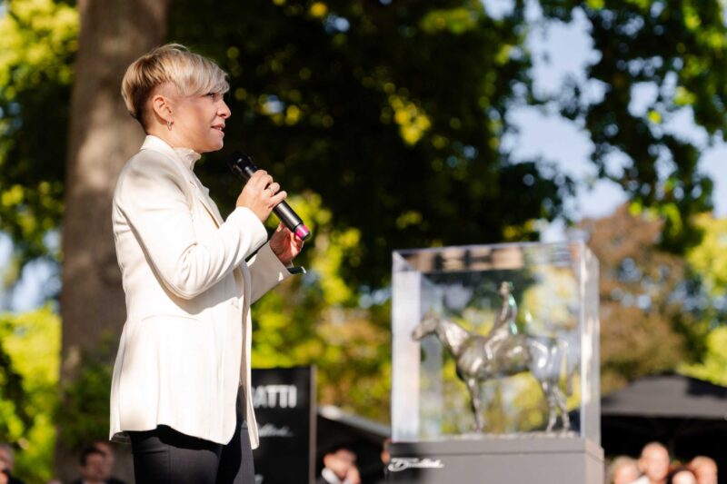 A person in a white blazer speaks into a microphone outdoors beside a glass case containing a horse and rider sculpture, as the 1-of-1 Bugatti Brouillard makes its European debut at Wheels Mariënwaerdt.
