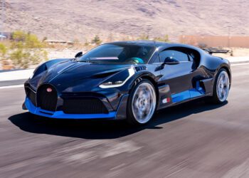 A blue carbon Bugatti Divo speeds along a paved road, set against a mountainous desert landscape, and is now for sale.