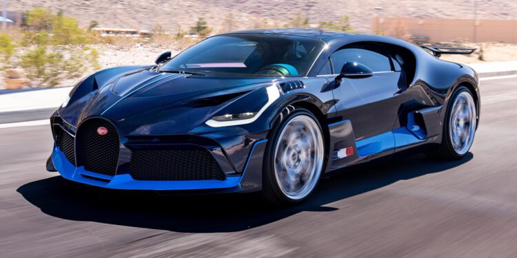 A blue carbon Bugatti Divo speeds along a paved road, set against a mountainous desert landscape, and is now for sale.