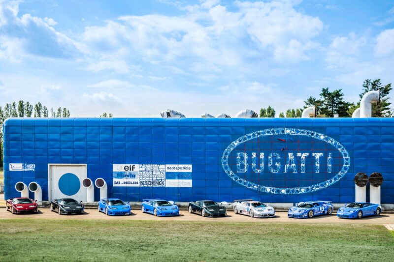 A row of sports cars is parked in front of a large blue Bugatti-branded building, where '90s hypercar royalty, the 1-of-39 Bugatti EB110 SS, hits the market under a partly cloudy sky.