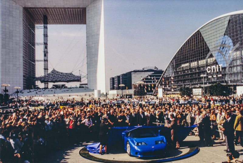 A crowd gathers around a rare '90s hypercar royalty: the 1-of-39 Bugatti EB110 SS, on display near the Grande Arche and CNIT building in Paris, with modern architecture visible in the background.
