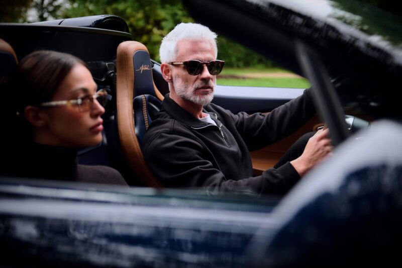 An older man with white hair and a woman with brown hair sit in a convertible car, both wearing stylish Tourbillon-Inspired Eyewear sunglasses and looking forward.